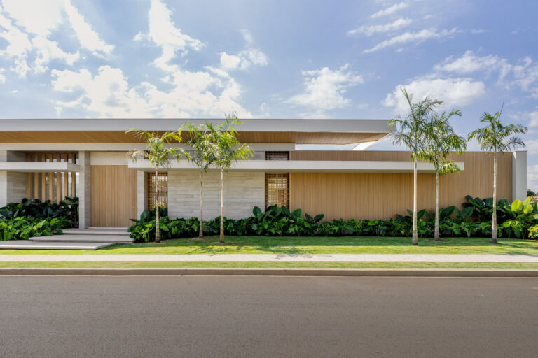 Fotografia de arquitetura residencial contemporânea mostrando a fachada térrea de linhas horizontais marcantes. A composição destaca o contraste entre as paredes de concreto aparente e os grandes painéis ripados de madeira clara. À frente, um paisagismo tropical com palmeiras e plantas de folhas largas (como costela-de-adão e guaimbê) emoldura a calçada e a rua asfáltica sob céu azul com nuvens.