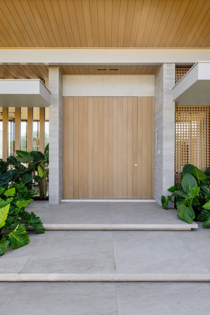 Alinhamento simétrico da entrada principal de uma residência de alto padrão. Uma imponente porta pivotante de madeira ripada clara é emoldurada por pilares de concreto aparente e marquise branca geométrica. O piso em grandes formatos de porcelanato cinza claro leva até o hall, ladeado por folhagens tropicais nas duas extremidades.